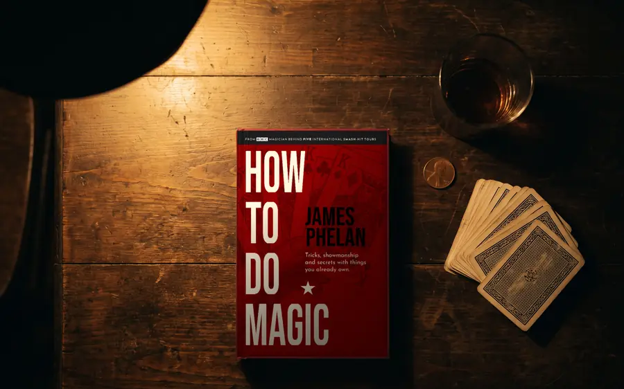 How to do Magic hardback on a dark wooden table in lamplight, beside a deck of cards, a coin and a glass